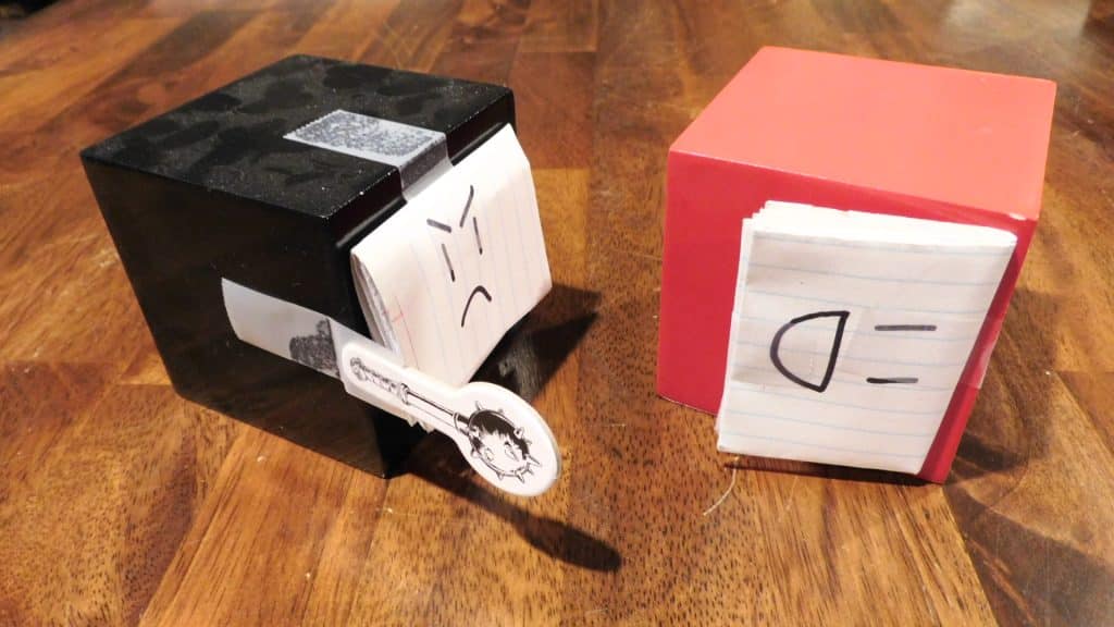 A black cube with a face taped to the front and a mace token taped to the side. Also, a red cube with a face taped to the front tipped over on its side.