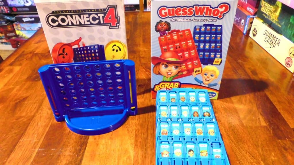 The boxes for Connect 4 Grab & Go and Guess Who? Grab & Go and their components in front of them on a table.