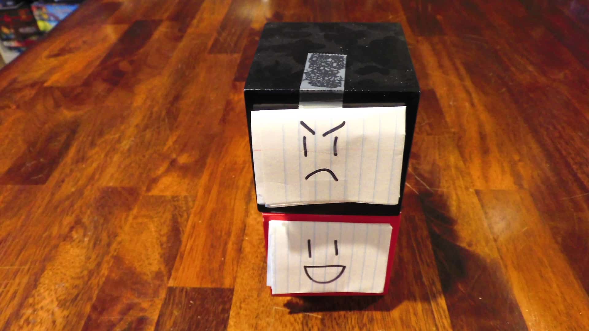 A black cube sitting on top of a red cube, each with a face on a piece of paper taped to their fronts.