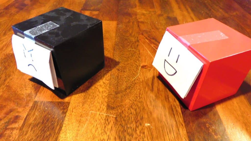 A red cube with a happy face on it and a black cube with an angry face on it facing the same way.
