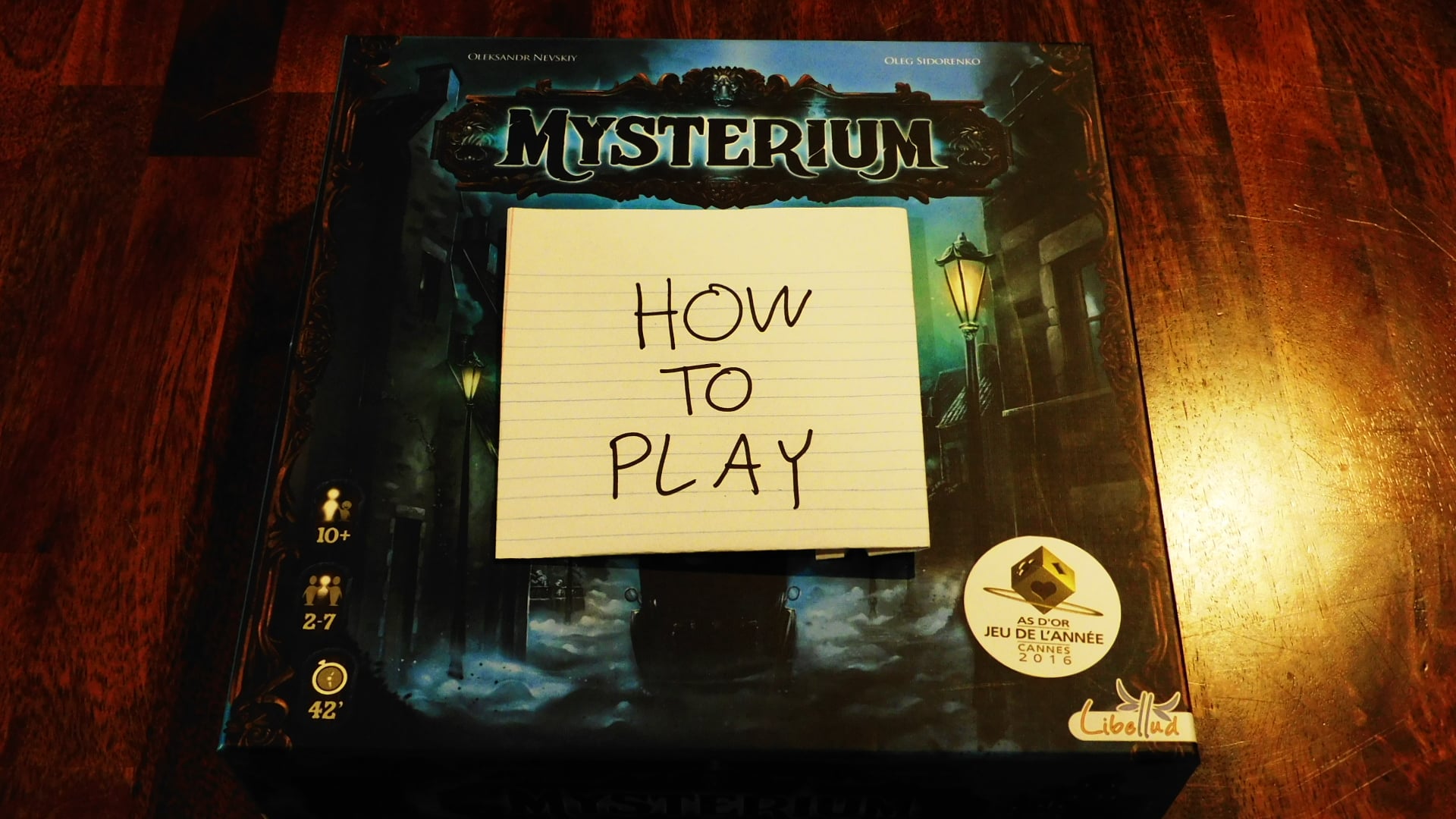 The Mysterium game box with "How To Play" written on a piece of paper on it.